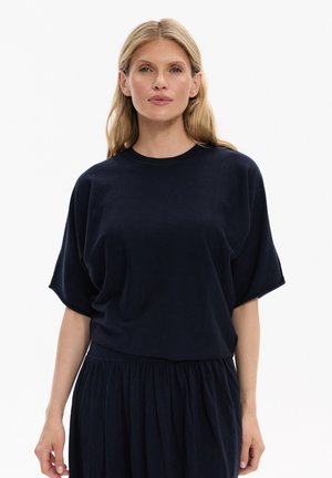 Blondynka ubrana w luźną czarną bluzkę z krótkim rękawem i dobraną czarną marszczoną spódnicę na białym tle.