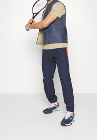 Marinblå quiltad väst över beige t-shirt, matchad med marinblå joggingbyxor med röd sidodetalj. Modell som håller en tennisracket.