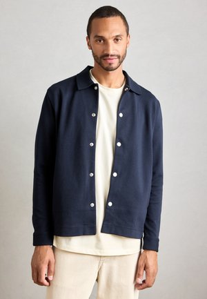 Man wearing a navy button-up jacket over a beige shirt and light beige pants, standing against a plain light gray background.