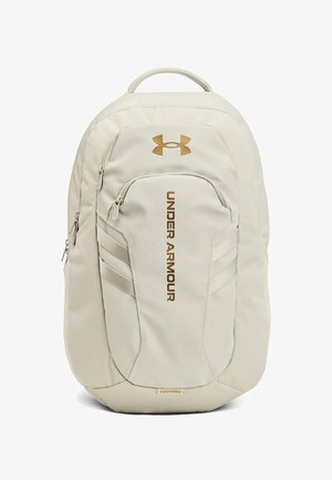 Jasnobeżowy plecak Under Armour z wieloma kieszeniami na suwak oraz logo i nazwą marki w brązowym kolorze na przednim panelu.
