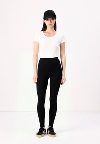 Zwarte leggings met een hoge taille, gecombineerd met een witte top met korte mouwen en een zwarte pet. De schoenen hebben een zwart- en beige ontwerp.