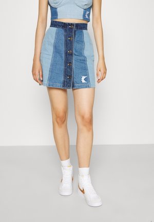 Femme portant une jupe en denim bleu bicolore avec des boutons sur le devant, associée à des baskets blanches montantes et des chaussettes blanches, debout sur un sol gris.