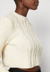 Krämfärgad stickad tröja med kabelmönster, så kallad cropped design, ribbad nederkant och rund halsringning. Strukturerad tyg med mjuk yta.