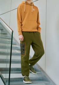 Felpa arancione con coulisse, logo bianco, pantaloni cargo verdi con tasca testurizzata e dettagli arancioni, abbinati a sneakers marroni e bianche.