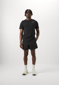 T-shirt Nike nera a maniche corte, pantaloni corti neri con texture e sneakers bianche con accenti gialli. Figura in piedi su uno sfondo chiaro.