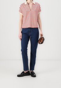 Červeno-bílý pruhovaný top s krátkým rukávem a výstřihem do V; v kombinaci se tmavě modrými džíny rovného střihu a černými mokasíny; drží hnědou kabelku.