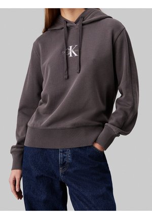 Un hoodie gris en tissu doux, avec une poche avant, une capuche avec cordon de serrage et un logo blanc. Associé à un jean bleu foncé.