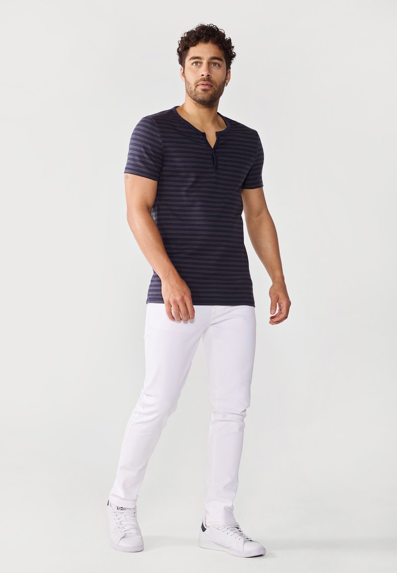 Camisa de rayas azul marino de manga corta con cuello abotonado, combinada con jeans blancos de corte ajustado y zapatillas blancas.