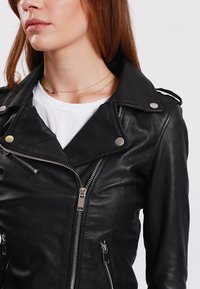 Giacca da moto in pelle nera con zip frontale, collare con bottoni a pressione e dettagli in metallo argentato. Texture liscia con due tasche laterali.