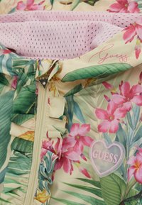 Giacca a zip floreale con fiori rosa e gialli su uno sfondo chiaro, dettagli in foglia verde, rifiniture a volant e un logo a forma di cuore trasparente.