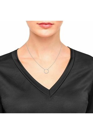 Gros plan d'une femme portant un délicat collier en chaîne argentée avec un petit pendentif circulaire texturé, vêtue d'un haut noir à col en V.