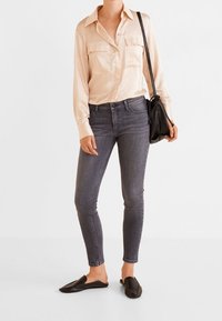 Camisa de seda beige con dos bolsillos en el pecho, combinada con pantalones ajustados gris oscuro y zapatos negros tipo slip-on. Incluye un bolso cruzado negro sencillo.