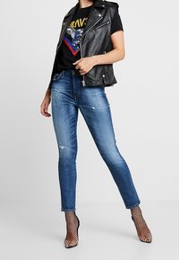 Černá kožená bezrukávová bunda přes černé tričko s potiskem, doplněná o světle modré úzké džíny s vysoko vytepanými detaily a černé síťované jehly.