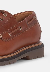 Chaussure en cuir marron avec un bout arrondi, des œillets en métal et une semelle en caoutchouc robuste. Elle possède un talon empilé et des détails de couture le long de l'empeigne.