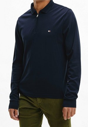 Mężczyzna ubrany w granatową koszulę polo z długim rękawem, z suwakiem i małym logo na piersi, połączoną z oliwkowymi spodniami.