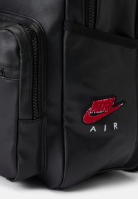 Crni kožni ruksak s patentnim zatvaračima, teksturiranom površinom i crvenim izvezenim Nike logom ispod kojeg piše "AIR".