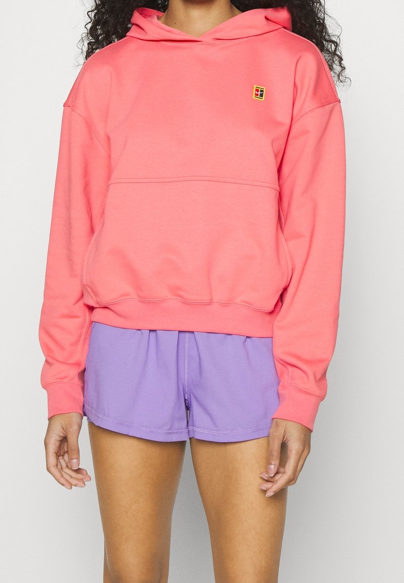 Femme portant un sweat à capuche rose corail avec un petit logo et un short de sport lavande, debout devant un fond clair uni.