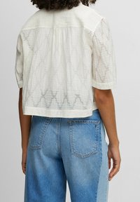 Kraftigt beskuren vit texturerad skjorta med korta ärmar, med vertikala ränder och ett subtilt vågmönster längs fållar, tillsammans med ljusblå jeans.