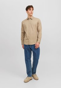 Tan long-sleeve button-up shirt with a smooth texture, paired with loose blue jeans and beige shoes, standing against a plain background.