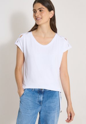 Femme souriante aux cheveux bruns portant un haut blanc à manches courtes avec des découpes aux épaules et un jean bleu clair, debout les mains dans les poches.
