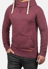 Man i en vinröd hoodie med krämfärgade dragsnören och frontficka, i kombination med mörkgrå jeans, står med en hand i fickan.