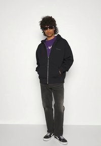 Sudadera negra con cremallera y bolsillo canguro, llevada sobre una camiseta gráfica morada, combinada con jeans oscuros y zapatillas negras.