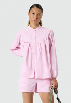 Vrouw draagt een lichtroze, lange mouwen blouse met verticale strepen en bijpassende korte broek, met een groene camo-patroon riem in haar hand.