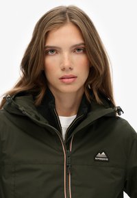 Ciemnozielona wodoodporna kurtka z kapturem, zapinana na zamek, z kontrastowymi pomarańczowymi akcentami. Na piersi znajduje się naszywka z logo marki.