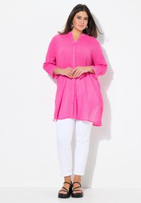 Fuchsia overhemd met knopen en een gestructureerde afwerking, voorzien van een kraag, driekwart mouwen en een losse pasvorm. Gepaard met witte broek en zwarte sandalen.