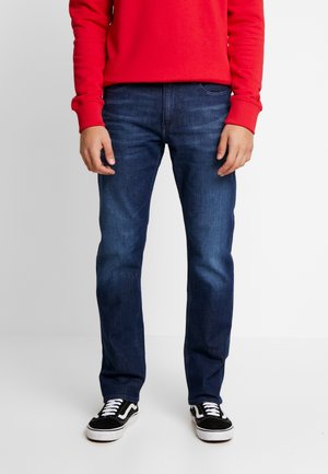 Homme portant un jean bleu foncé, une chemise rouge à manches longues et des baskets noires et blanches, debout devant un fond uni.