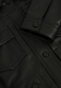 Schwarze Lederjacke mit Druckknopfmanschetten und aufgesetzten Taschen. Glatte Textur mit sichtbaren Nähdetails und rautenförmigen gesteppten Einsätzen.