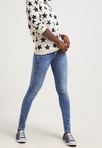 Weißer Kapuzenpullover mit dunkelblauem Sternmuster, kombiniert mit blauen Skinny-Jeans und dunkelblauen Sneakers mit weißen Schnürsenkeln. Lässiges Outfit in einem hellen Ambiente.