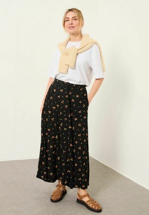 Čierna kvetinová sukňa s červenými a žltými vzormi, biele tričko a krémový pletený sveter zviazaný okolo krku. Hnedé sandále s remienkami dopĺňajú vzhľad.