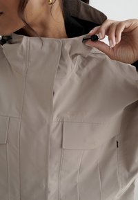 Vrouw die de kraag van een beige jas met zwarte koordstopper verstelt, zichtbare oorbel en dichtgeknoopte borstzak.