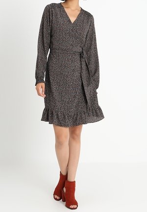 Kobieta ubrana w długą sukienkę kopertową z długim rękawem, dekoltem w serek, sięgającą do kolan, w kwiatowy wzór, z falbaną na dole oraz czerwonymi, odkrytymi, na wysokim obcasie, botkami do kostki.