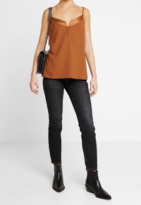 Kamelkově zbarvený top s výstřihem do V, s knoflíky vpředu a tenkými ramínky. V tomto outfitu jsou kombinovány s tmavými skinny džíny a černými kotníkovými botami.