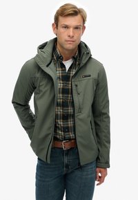 Uomo con giacca verde oliva con cappuccio e zip aperta sopra camicia a quadri verde e giallo, jeans blu e cintura marrone, in piedi davanti a uno sfondo bianco.