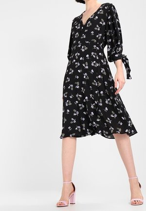 Kobieta ubrana w czarną, kwiecistą sukienkę sięgającą kolan z bufkami związanymi na mankietach, do tego różowe sandały na wysokim obcasie.