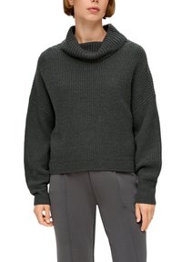 Frau trägt einen dunkelgrauen, grob gestrickten Pullover mit weit gefaltetem Kragen und grauen, eng anliegenden Hosen, steht vor einem weißen Hintergrund.