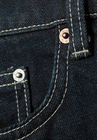 Dark denim fabric features white stitching, copper and silver rivets on a front pocket, with a smooth and textured finish.