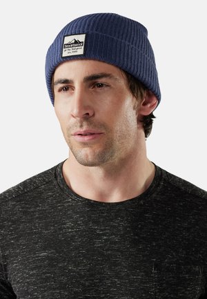 Gorro de punto acanalado en color azul marino con un parche de logo en el frente, llevado por una persona que viste una camiseta de cuello redondo oscura y texturizada. Enfoque en la forma ajustada y el color del gorro.