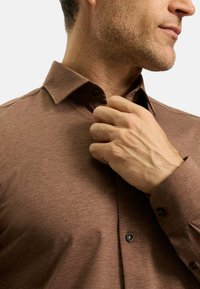 Braunes Langarmhemd aus strukturiertem Stoff, mit einem spitzen Kragen und schwarzen Knöpfen. Das Hemd ist am Kragen teilweise zugeknöpft.