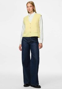 Žuta pletena prsluk s V-izrezom, prednjim gumbima i dvije džepove, uparen s tamnoplavim trapericama širokih nogavica i crnom obućom.