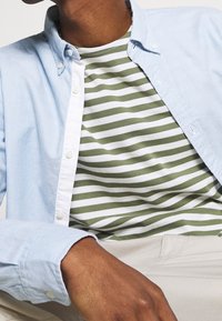 Personne portant une chemise bleu clair à boutons ouverte sur un t-shirt rayé vert et blanc, associée à un pantalon beige.