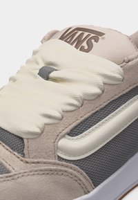 Εγγύς πλάνο μπεζ και γκρι παπουτσιού Vans με κορδόνια εκρού και υφασμάτινη πλέξη, εμφανίζοντας το λογότυπο Vans στη γλώσσα.