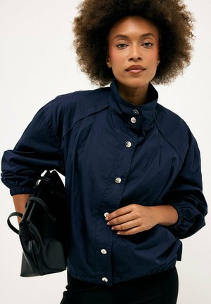 Donna con capelli ricci che indossa una giacca blu navy abbottonata, tiene una borsa in pelle nera con una mano alla vita.