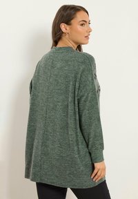 Femme portant un pull vert ample à manches longues avec une couture dans le dos, se tenant contre un mur blanc uni, de profil.