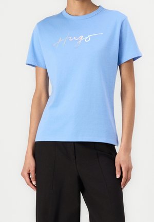 Osoba w jasnoniebieskiej koszulce z krótkim rękawem z metalicznym napisem „Hugo” oraz czarnych spodniach, stojąca na jednolitym białym tle.