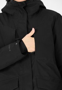 Svart vattentät jacka med hög krage, dragkedjor och externa fickor. Den har en tumhål-design och mjukt foder.