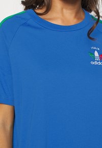 Modré bavlněné tričko se zelenými akcenty na ramenou, zdobené vyšívaným nápisem "Italia" a vícebarevným logem ve tvaru trojlístku. Kulatý výstřih a krátké rukávy.